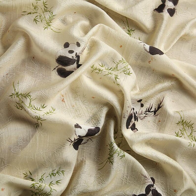 Pu&ograve; includere: Un tessuto color crema con un motivo ripetuto di panda bianchi e neri e steli di bamb&ugrave; verdi. Il materiale presenta una superficie sottilmente strutturata con pieghe e drappeggi visibili, che mettono in risalto le illustrazioni animali.