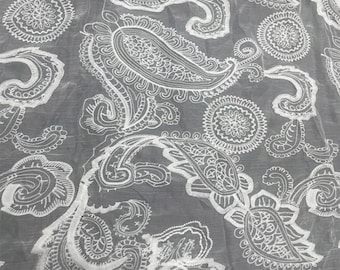 May include: Close-up of a sheer fabric with a white paisley pattern. The design features detailed, swirling floral and teardrop shapes. The fabric has a soft, flowing appearance, with the white pattern standing out against the gray background.