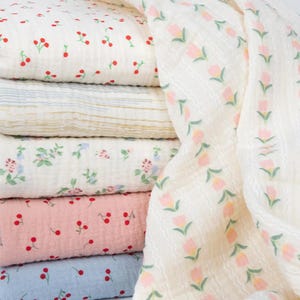 May include: A stack of soft, patterned fabric. The patterns include cherries, floral designs, and stripes in various colors like white, pink, and blue. A separate piece of fabric with a tulip pattern is draped on the side.