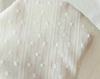 May include: A sheer off-white fabric featuring a delicate texture with vertical stripes and small, raised tufted dots scattered across the surface. The material is lightweight and semi-transparent, showcasing a consistent pattern of woven lines.