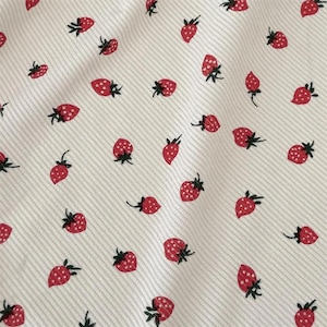 May include: Close-up of white fabric featuring a repeating pattern of red strawberries with green stems. The strawberries are dotted and scattered across the fabric, which has a subtle striped texture.
