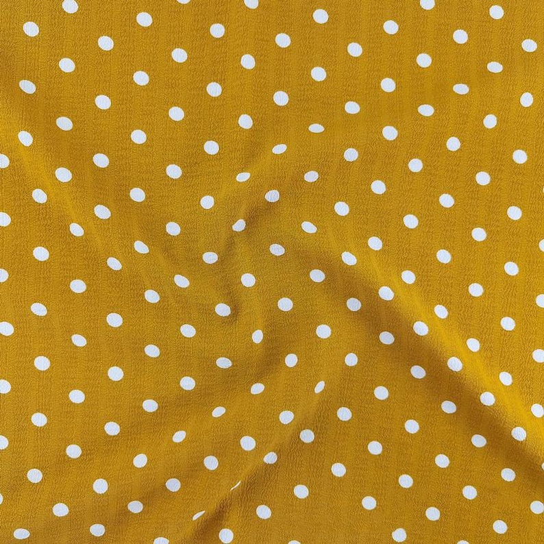 May include: A mustard yellow fabric featuring a repeating pattern of small, evenly spaced white polka dots. The material has a soft, slightly textured surface with gentle folds that create subtle shadows across the dotted design.