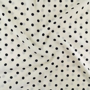 May include: A lightweight fabric featuring a repeating pattern of solid black polka dots evenly spaced across an off-white background. The material displays a soft, slightly crinkled texture with natural folds that create subtle shadows.