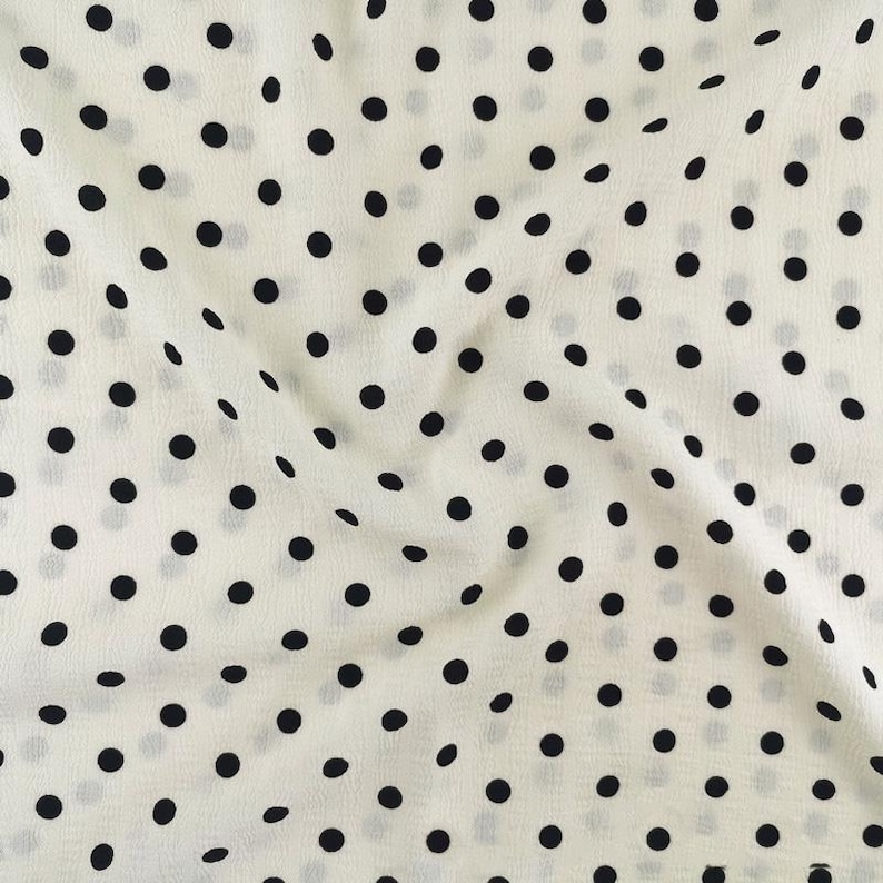 May include: A lightweight fabric featuring a repeating pattern of solid black polka dots evenly spaced across an off-white background. The material displays a soft, slightly crinkled texture with natural folds that create subtle shadows.