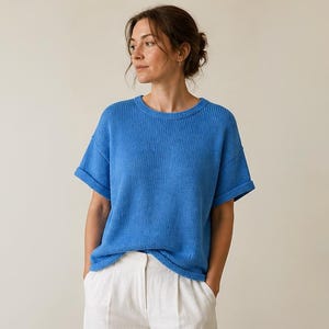 May include: A blue short-sleeved top with a ribbed texture. The top has a crew neck and rolled-up sleeves. The model is wearing white trousers. The top is a vibrant shade of blue.