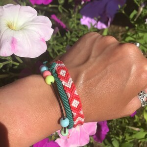 May include: A close-up of a wrist wearing three bracelets. The bracelets are made of colourful string and beads. The top bracelet is red and white with a diamond pattern. The middle bracelet is green and the bottom bracelet is black with colourful beads.