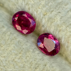 May include: Two oval-cut, faceted rubies. The gemstones are a deep, saturated red with internal reflections. The rubies are set against a soft, off-white, textured background.