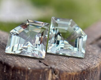 May include: Two faceted, light green gemstones with a geometric cut. The gems are displayed on a weathered, brown wooden surface, with a blurred green background. The gemstones have a clear, crystalline appearance.