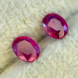 Könnte beinhalten: Zwei oval geschliffene, facettierte Rubine. Die Edelsteine sind ein tiefes, leuchtendes Rot mit violetten Farbtönen. Die Rubine werden auf einer strukturierten, cremefarbenen Oberfläche präsentiert, die ihre Brillanz und Klarheit hervorhebt. Ideal für die Schmuckherstellung.