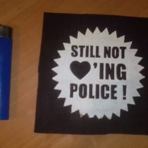 May include: A black patch with a white circle and the text "STILL NOT 'ING POLICE!" in black lettering. A black heart is in the center of the circle.