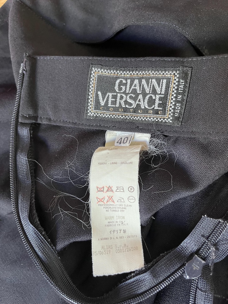 May include: Close-up of a black garment with a zipper and a label. The label reads "GIANNI VERSACE COUTURE" and "MADE IN ITALY". A size tag shows the number "40". The garment has care instructions and other text.
