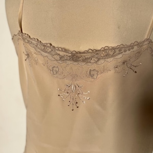May include: A beige camisole with delicate spaghetti straps. The top features a scalloped lace trim along the neckline and intricate embroidered floral detailing on the back. The fabric appears to be a soft, flowing material.