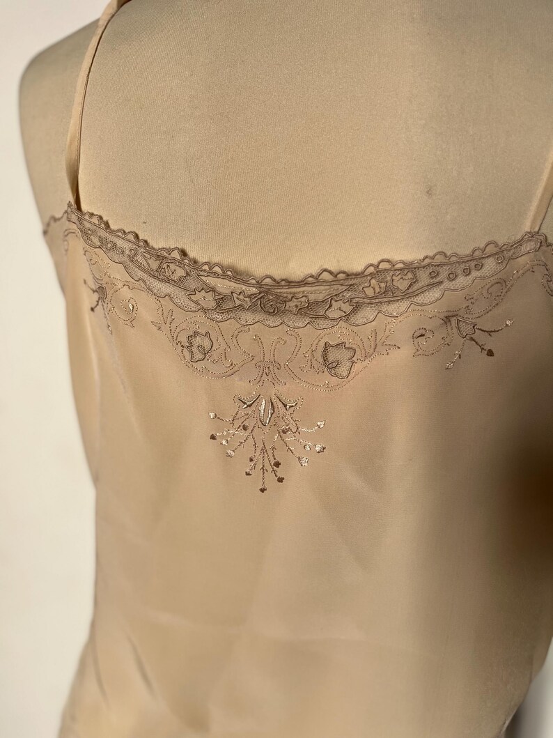 May include: A beige camisole with delicate spaghetti straps. The top features a scalloped lace trim along the neckline and intricate embroidered floral detailing on the back. The fabric appears to be a soft, flowing material.