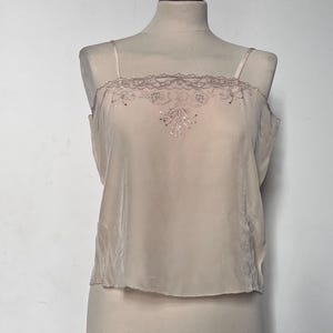 May include: A light beige camisole top with delicate spaghetti straps. The top features a decorative lace trim along the neckline and a subtle embroidered design on the front. The garment is displayed on a mannequin.