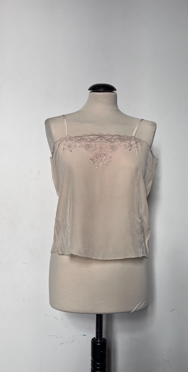 May include: A light beige camisole top with delicate spaghetti straps. The top features a decorative lace trim along the neckline and a subtle embroidered design on the front. The garment is displayed on a mannequin.