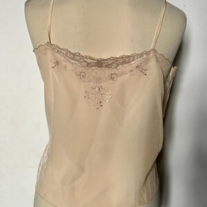 May include: A light peach-colored camisole with delicate spaghetti straps. The top features a lace trim along the neckline and a subtle embroidered design on the back. The fabric appears to be a soft, flowing material.