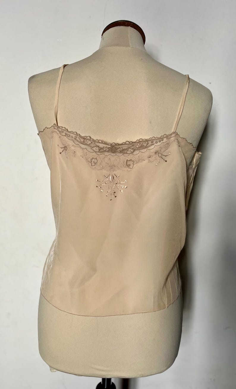 May include: A light peach-colored camisole with delicate spaghetti straps. The top features a lace trim along the neckline and a subtle embroidered design on the back. The fabric appears to be a soft, flowing material.