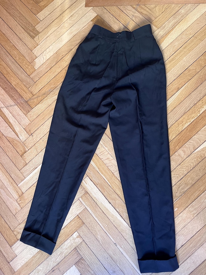 May include: Black trousers with a high waist and a tailored fit. The pants have a classic design with pleats at the front and cuffs at the hem. The dark color and simple style make them versatile for various outfits.