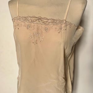 May include: A beige camisole top with thin straps, displayed on a mannequin. The top features a delicate lace trim along the neckline and a floral embroidery design on the front. The fabric appears to be a soft, silky material.