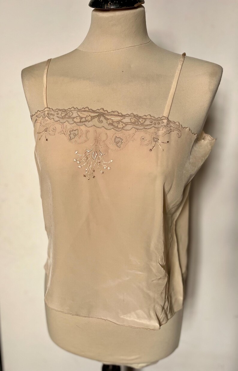 May include: A beige camisole top with thin straps, displayed on a mannequin. The top features a delicate lace trim along the neckline and a floral embroidery design on the front. The fabric appears to be a soft, silky material.