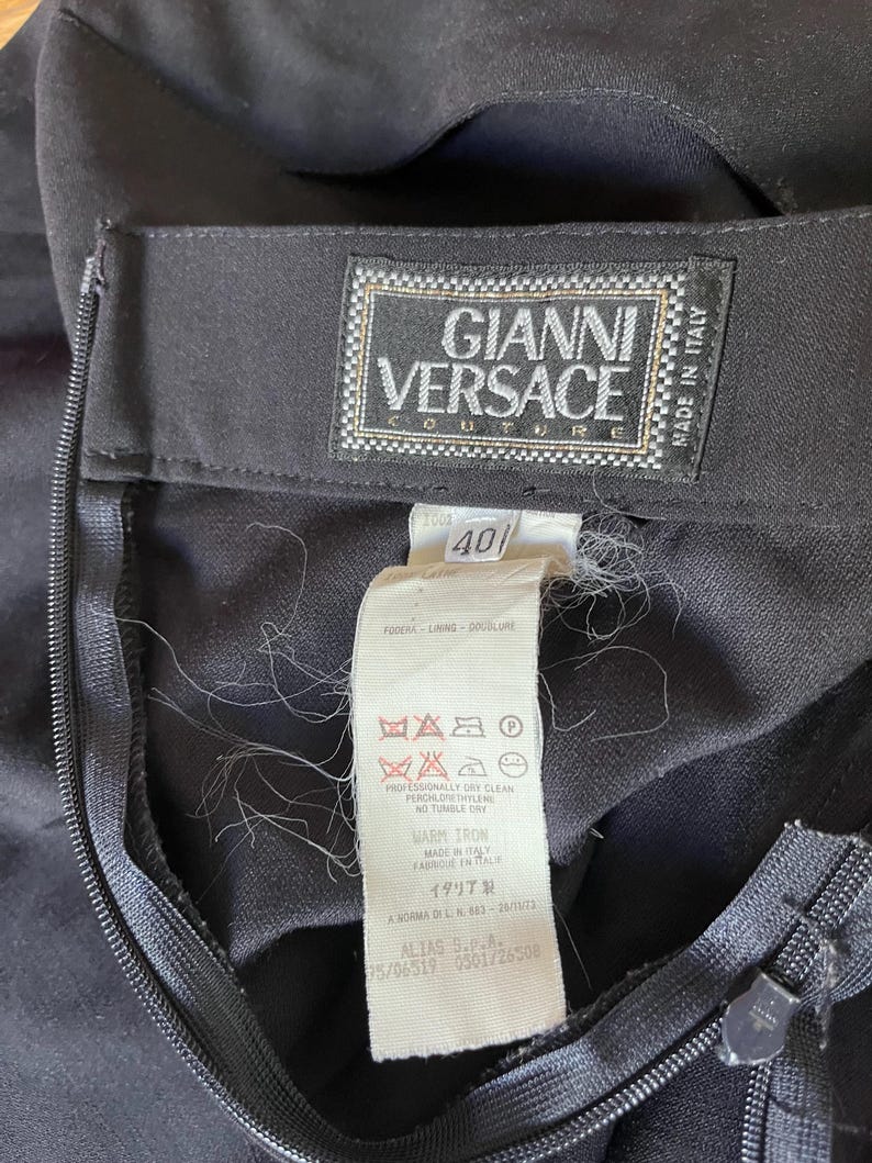 May include: Close-up of a black garment with a zipper and a Gianni Versace Couture label. The label includes the text "Made in Italy" and size 40. Additional care instructions are visible.