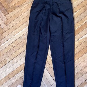 May include: Dark navy blue high-waisted trousers with a pleated front and cuffed hems. The pants are made of a smooth, solid fabric. The trousers are displayed on a wooden floor.