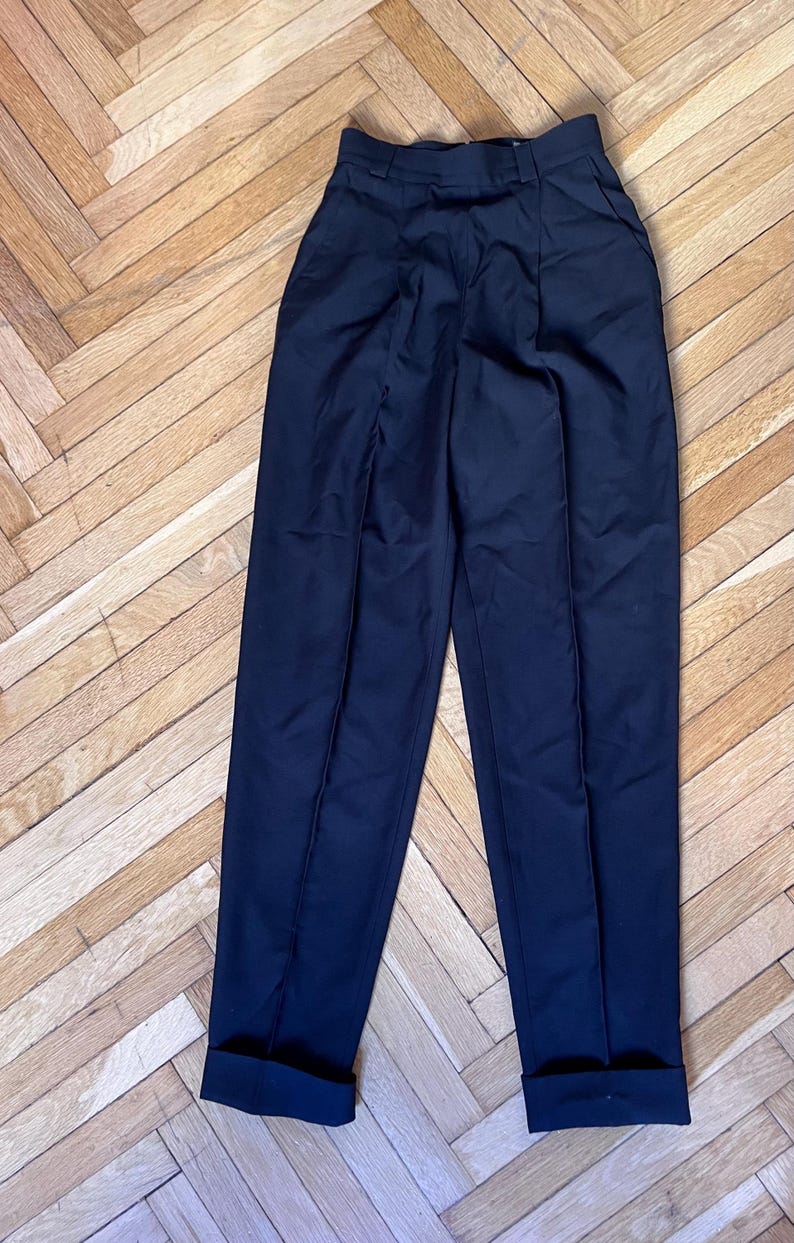 May include: Dark navy blue high-waisted trousers with a pleated front and cuffed hems. The pants are made of a smooth, solid fabric. The trousers are displayed on a wooden floor.
