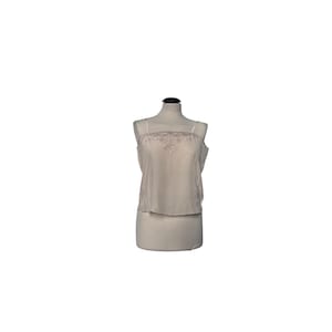 May include: A beige camisole top with thin spaghetti straps and a lace trim along the neckline. The top is displayed on a mannequin. The fabric appears to be a soft, flowing material. The camisole has a simple, elegant design.