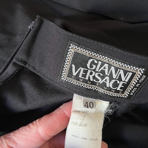 May include: Close-up of a black garment with a label that reads "GIANNI VERSACE". The label is white with gold trim and the size tag reads "40". The fabric is 100% wool.