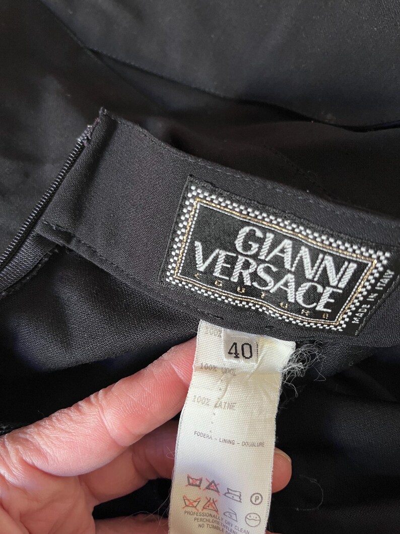 May include: Close-up of a black garment with a label that reads "GIANNI VERSACE". The label is white with gold trim and the size tag reads "40". The fabric is 100% wool.