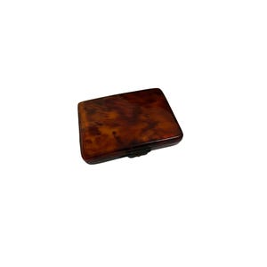 May include: A rectangular, brown tortoiseshell cigarette case with a hinged lid and a dark metal clasp. The case has a glossy finish and a marbled pattern, suggesting a vintage or antique style.