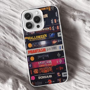 May include: A clear phone case with a design of stacked VHS tapes. The tapes feature titles such as "Halloween," "Phantasm," and "Scream." The case has a white border and covers the phone's back and sides.