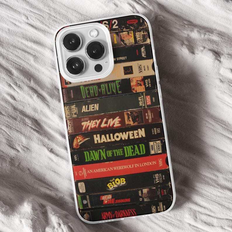 May include: A phone case displaying a collage of old VHS tapes, including titles such as "Dead Alive," "Alien," and "Halloween." The case has a transparent edge and a camera aperture. The design has a retro horror movie aesthetic.