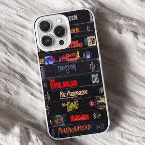 May include: A phone case displaying a collection of retro VHS tapes, including titles such as "The Thing" and "A Nightmare on Elm Street." The case has a clear border and a white camera module.