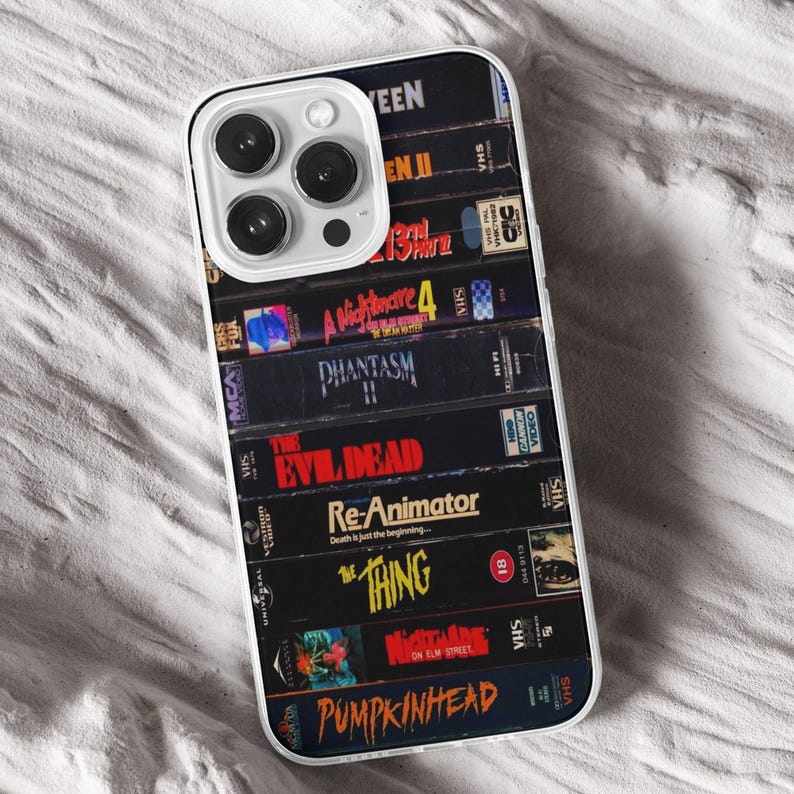 May include: A phone case displaying a collection of retro VHS tapes, including titles such as "The Thing" and "A Nightmare on Elm Street." The case has a clear border and a white camera module.