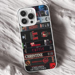 May include: A clear phone case displaying a collection of VHS tapes, including "Misery", "The Shining", and "Carrie". The case has a white border and a camera cutout.