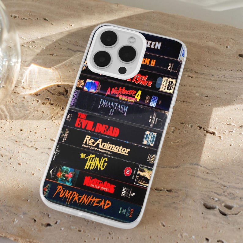 May include: A phone case displaying a collection of VHS tapes with film titles including "The Evil Dead" and "A Nightmare on Elm Street." The case has a clear border and a multi-lens camera cutout.