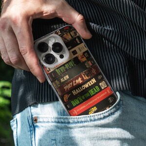 May include: A clear phone case displaying a collection of retro VHS tapes, including titles such as "Dead Alive," "Alien," and "Halloween." The case is partially visible in a pocket, evoking a nostalgic movie theme.