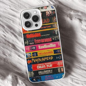 May include: A phone case with a design of stacked VHS tapes, including titles such as "Nightmare on Elm Street" and "Child's Play." The case has a clear body and a white border, with a camera aperture.