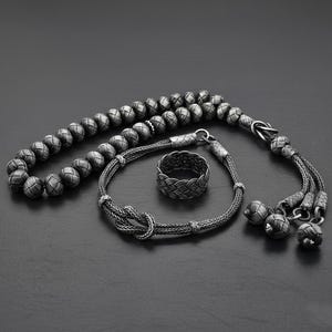 May include: A collection of silver-coloured jewellery, featuring a beaded strand, a bracelet with a knot design, and a woven ring. The items are displayed on a dark surface, highlighting the intricate details.