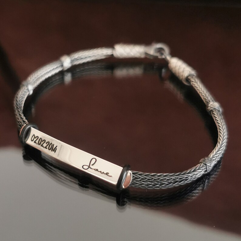 May include: A silver bracelet with a black braided cord and a silver rectangular tag engraved with the date "02022014" and the word "Love".