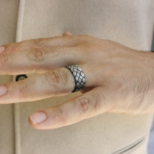 May include: A silver ring featuring a woven pattern. The ring is displayed on a finger, emphasizing the detailed design of intertwined metal strands. The ring's texture and design are the main focus, highlighting its craftsmanship.