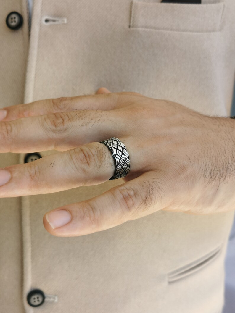 May include: A silver ring featuring a woven pattern. The ring is displayed on a finger, emphasizing the detailed design of intertwined metal strands. The ring's texture and design are the main focus, highlighting its craftsmanship.