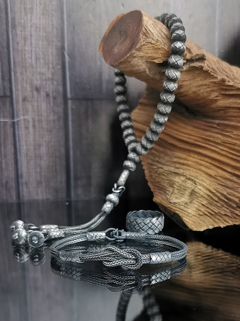 May include: A selection of silver jewellery, featuring a beaded necklace, a bracelet with a knot design, and a woven ring. The items are presented on a reflective surface with a wooden element in the background.