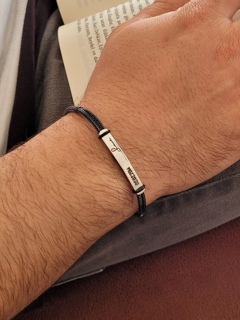May include: A black braided bracelet with a silver rectangular charm engraved with the date "02.02.2014" and a small infinity symbol.