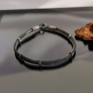 May include: A black braided silver bracelet with four silver knots. The bracelet is on a black reflective surface.