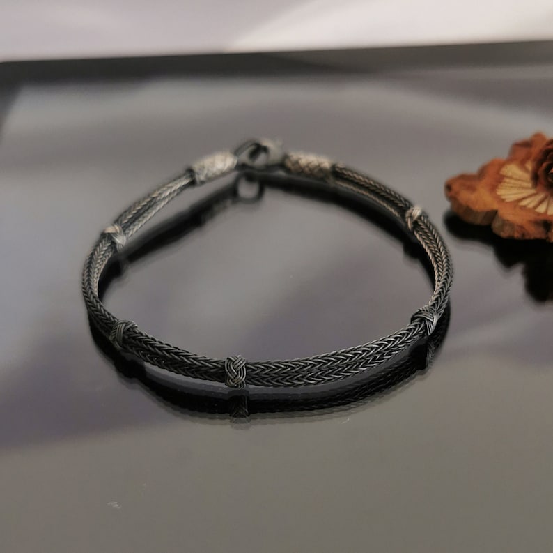 May include: A black braided silver bracelet with four silver knots. The bracelet is on a black reflective surface.