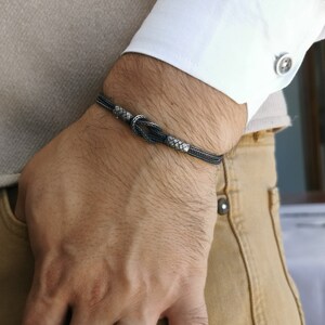 May include: A silver bracelet with a woven design and a knot detail is worn on a wrist. The bracelet is paired with a matching woven silver ring. The person is wearing a white shirt and tan trousers.