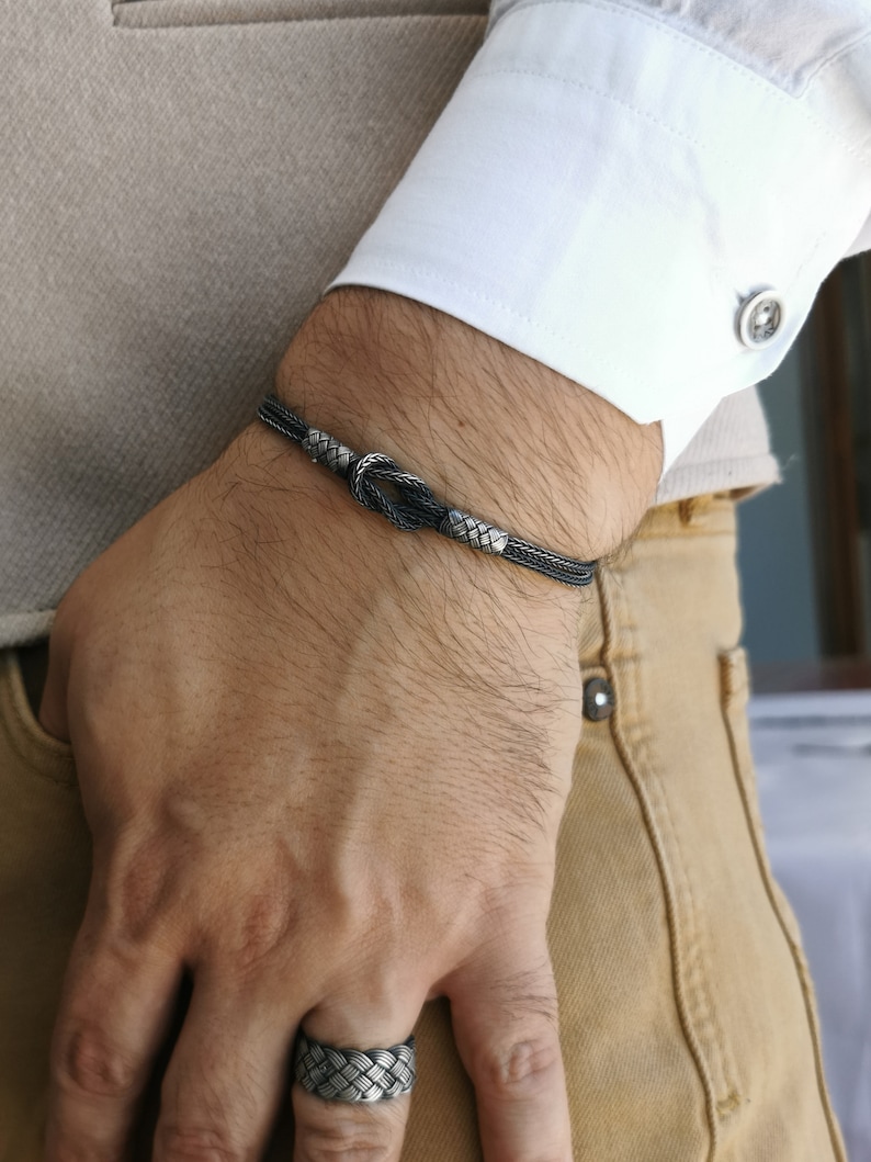May include: A silver bracelet with a woven design and a knot detail is worn on a wrist. The bracelet is paired with a matching woven silver ring. The person is wearing a white shirt and tan trousers.