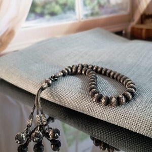May include: A silver-toned beaded necklace with a woven design. The necklace is resting on a grey textured surface. The necklace has a tassel of small, decorative silver balls.